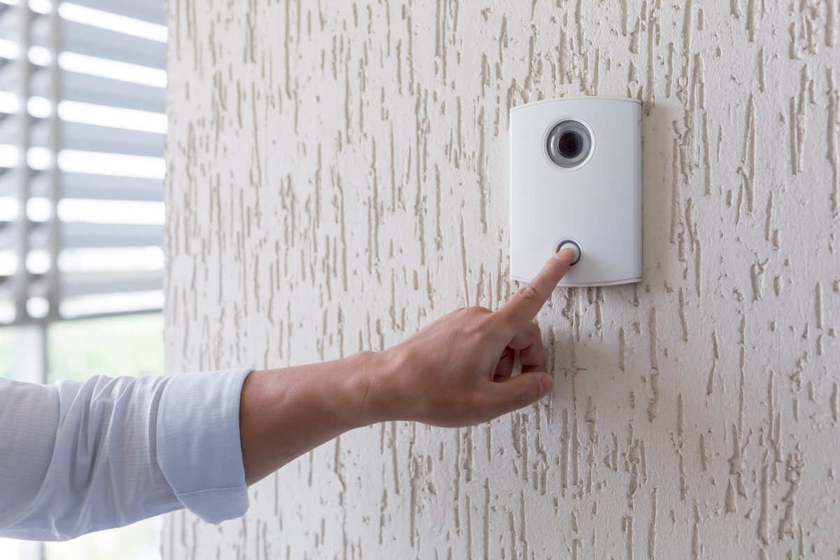A close-up of a hand pressing the button on a modern white video doorbell mounted on a textured exterior wall.
