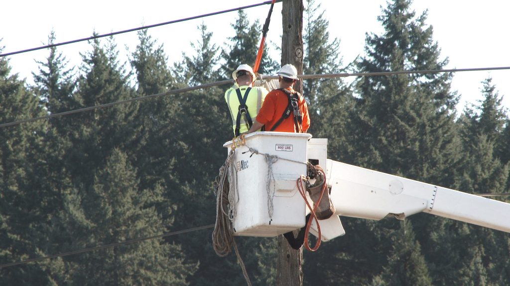 Two utility workers wearing hard hats and safety harnesses work from an elevated bucket beside a wooden utility pole, with power lines and evergreen trees in the background.